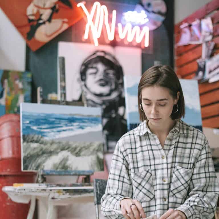 art student with art pieces behind her on wall as she works on a piece on her lap